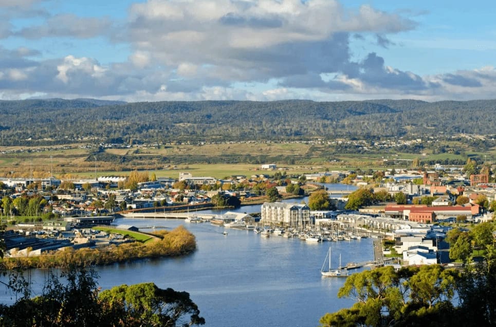 Coastal town with a river and marina, mountains in background.