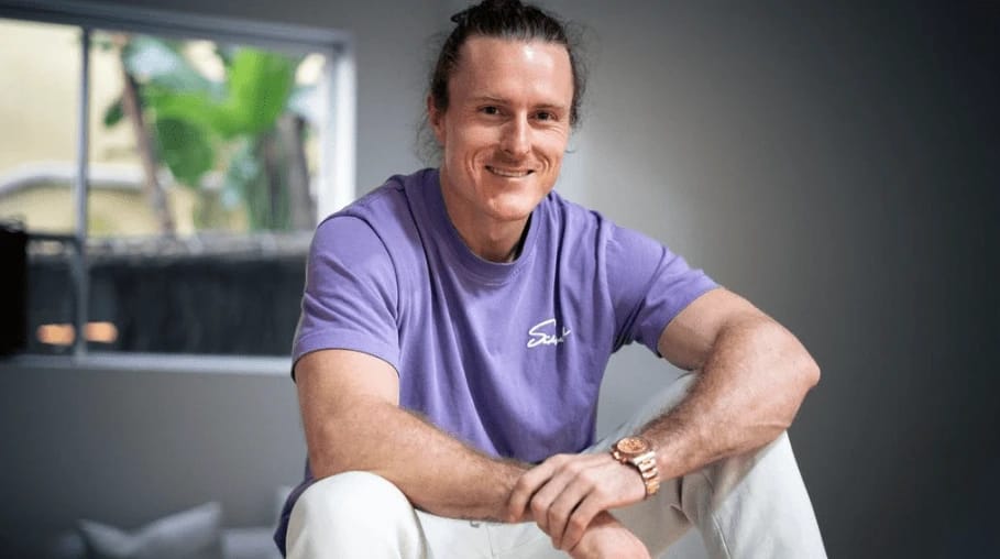 Man in purple t-shirt and white pants smiling.