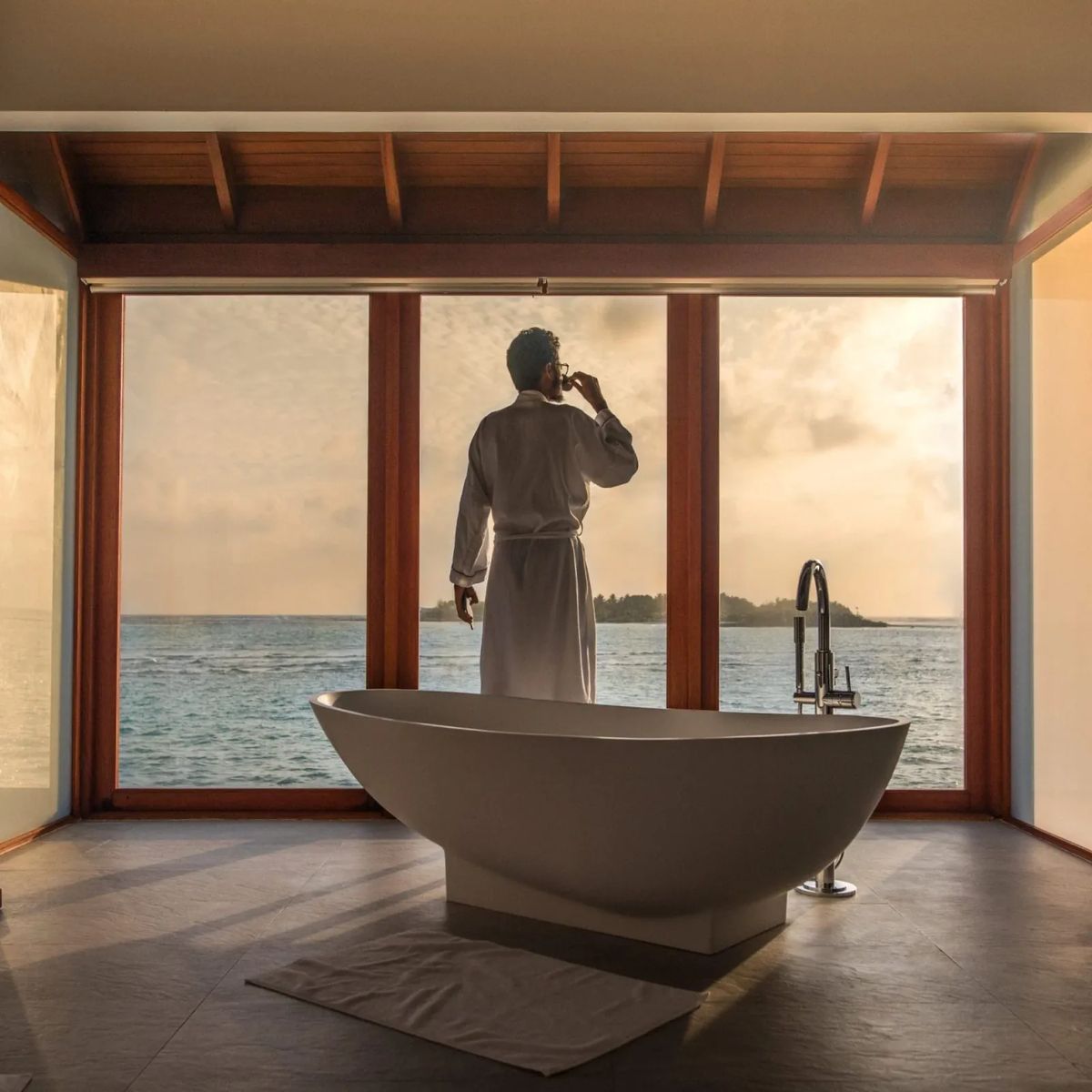 Man in robe drinks near tub facing ocean view.