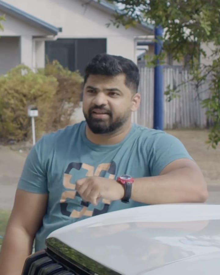 Manu, with short dark hair and a full beard, in a teal printed T-shirt, leaning on the bonnet of a white car in front of a house