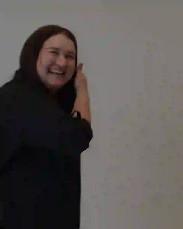 Dee, the local real estate agent, with long dark hair and wearing black, smiling in a real estate office