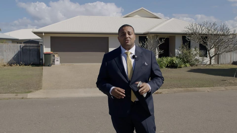 Arjun Paliwal in a navy suit and yellow tie standing on a street in front of a single-storey house with a double garage, a wheelie bin and a bare tree, under a blue sky