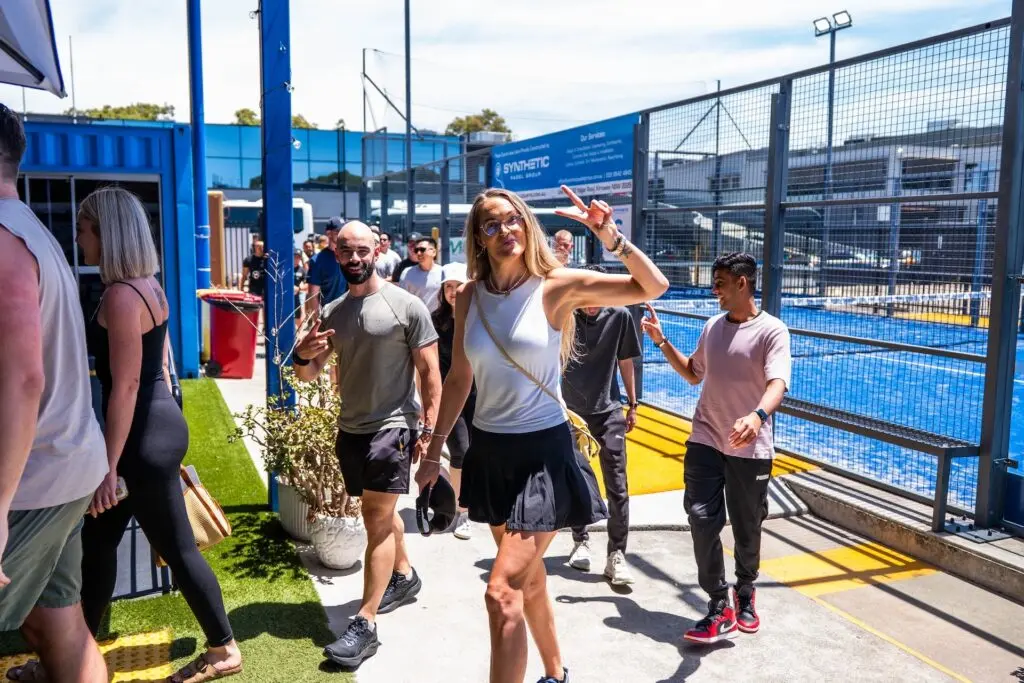 People walking by a padel court, a woman makes a peace sign.